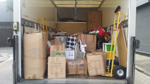 Inside the open rear of a commercial van, there is a large collection of discarded cardboard boxes and packaging materials, some flattened and others partially intact, with various printed labels and tape visible on their surfaces. The boxes occupy most of the space, stacked irregularly from front to back, with a few smaller boxes placed on top of larger ones. To the right side of the van, a yellow hand truck with black wheels leans against the boxes, and a small green planter with artificial flowers is positioned near the back wall. The background shows the van's interior walls, which are wooden and light-colored, with a dark cargo area above. The van appears to be parked on a paved driveway or street, with the neighbouring building's roller shutter door partially visible on the right. The scene suggests a recent rubbish removal operation by Commercial Waste Removal Chiswick, specialising in the collection and disposal of various waste items, including packaging debris typical in commercial or renovation settings in Chiswick and the surrounding postcode area.