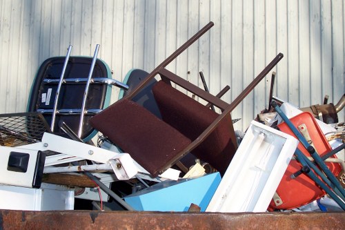 A pile of assorted waste objects stacked in an outdoor setting, including a brown fabric-upholstered chair with four wooden legs, a blue plastic container, a white-framed window, a red plastic container, and various metal objects such as a bicycle basket and a metal frame. The items are placed on a rusted metal surface with a corrugated metal wall in the background, suggesting an industrial or commercial area in Chiswick. The collection appears to consist of household and office rubbish awaiting removal, illustrating the type of clutter managed by Commercial Waste Removal Chiswick. The natural lighting highlights the different textures and colours of the materials, from the smooth plastics to the rough metal surfaces, providing a clear and detailed view of the waste collection ready for disposal or recycling.