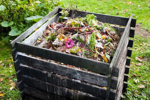 A black wooden compost bin filled with organic waste such as vegetable scraps, fallen leaves, and garden clippings, situated outdoors on a grassy area with some greenery and plants around it. The materials inside the bin display varying textures and colours, including brown composted matter, green leaves, and a few pink flowers. The bin is slightly weathered, with visible wood grain and some dirt on its surface, and is positioned on a patch of green lawn with small patches of soil and scattered leaves nearby. The overall scene indicates a typical garden compost setup, with natural daylight illuminating the area, aligning with waste management practices in residential or small commercial settings within the Chiswick area.