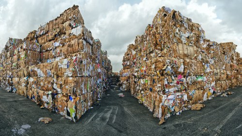Two large stacks of compressed cardboard and paper waste are positioned on either side of a central pathway on a paved outdoor surface. The waste materials, primarily consisting of brown corrugated cardboard and various types of paper, are tightly compacted into rectangular blocks, with some edges appearing rough and uneven. The stacks extend upward, dominating the scene against a cloudy sky overhead. Surrounding the stacks, the ground appears slightly worn, with a few scattered pieces of paper and small debris. In the background, there are no distinct structures, only the vast expanse of the sky, which is filled with grey clouds. The environment suggests an industrial or waste management setting, consistent with a rubbish removal operation, as handled by services like Commercial Waste Removal Chiswick. The scene emphasizes the scale of waste storage or disposal, highlighting the importance of professional rubbish removal for commercial clients in the local postcode area around Chiswick.